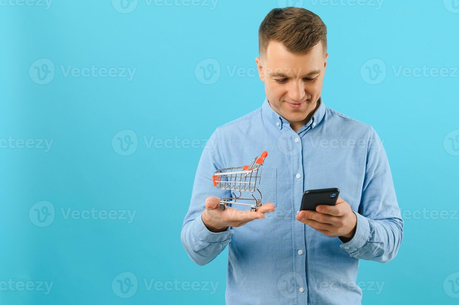 A young man holding a mini shopping cart and standing in front of a blue background in the studio. Sales concept. Place for text. photo