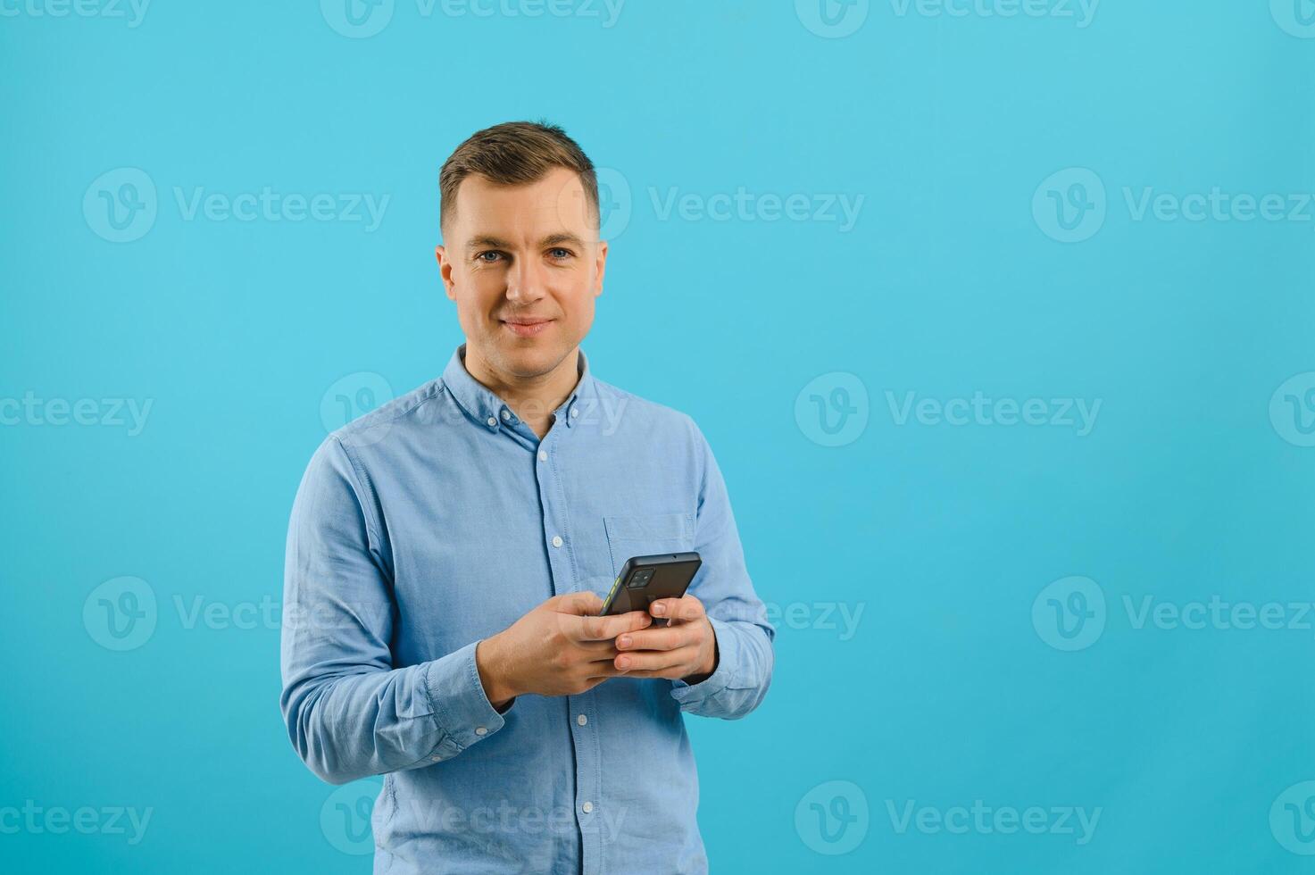 Photo of happy handsome man holding mobile phone in hand, texting. Young guy with big toothy smile posing on blue background.
