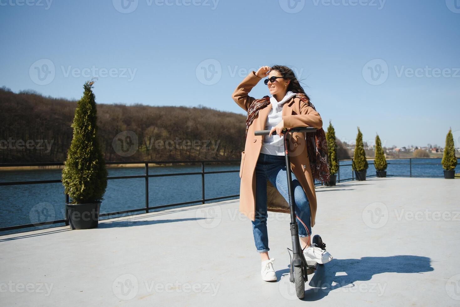 Young beautiful woman in a jacket smiles and rides an electric scooter to work along office buildings photo