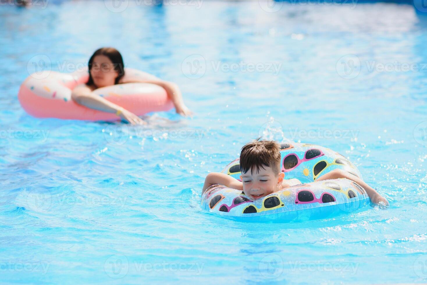 Happy family, young active mother and adorable curly little baby having fun in a swimming pool, child learning to swim in an inflatable toy ring, enjoying summer vacation at a tropical resort photo