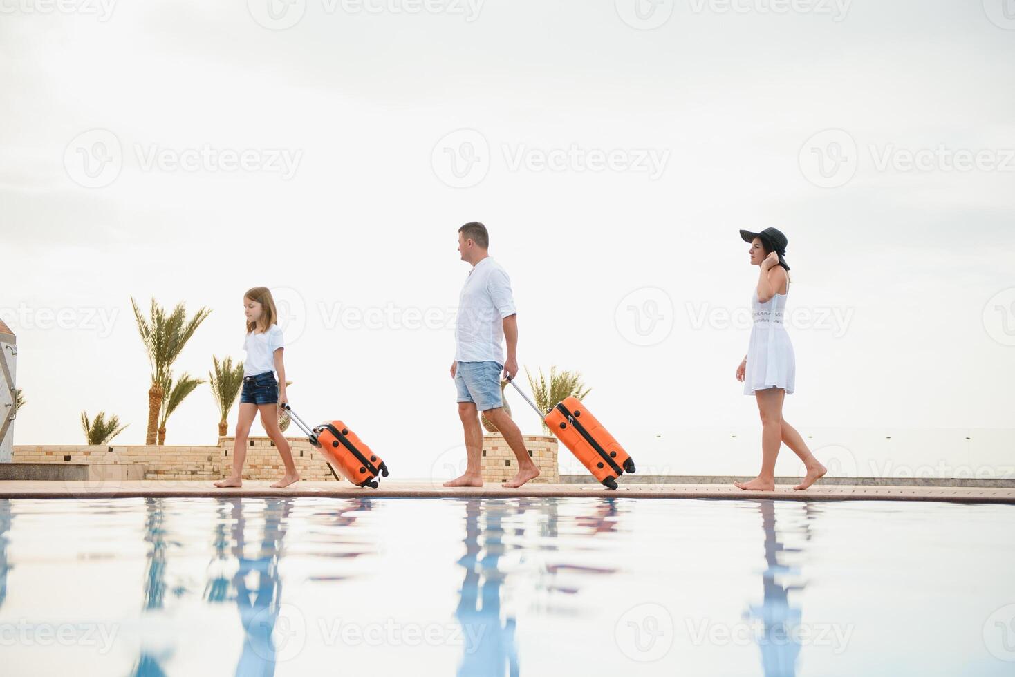 Young couple going to hotel through swimming pool upon arrival, looking for room, holding suitcases photo