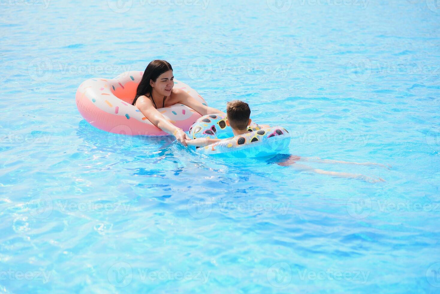 Happy family, young active mother and adorable curly little baby having fun in a swimming pool, child learning to swim in an inflatable toy ring, enjoying summer vacation at a tropical resort photo