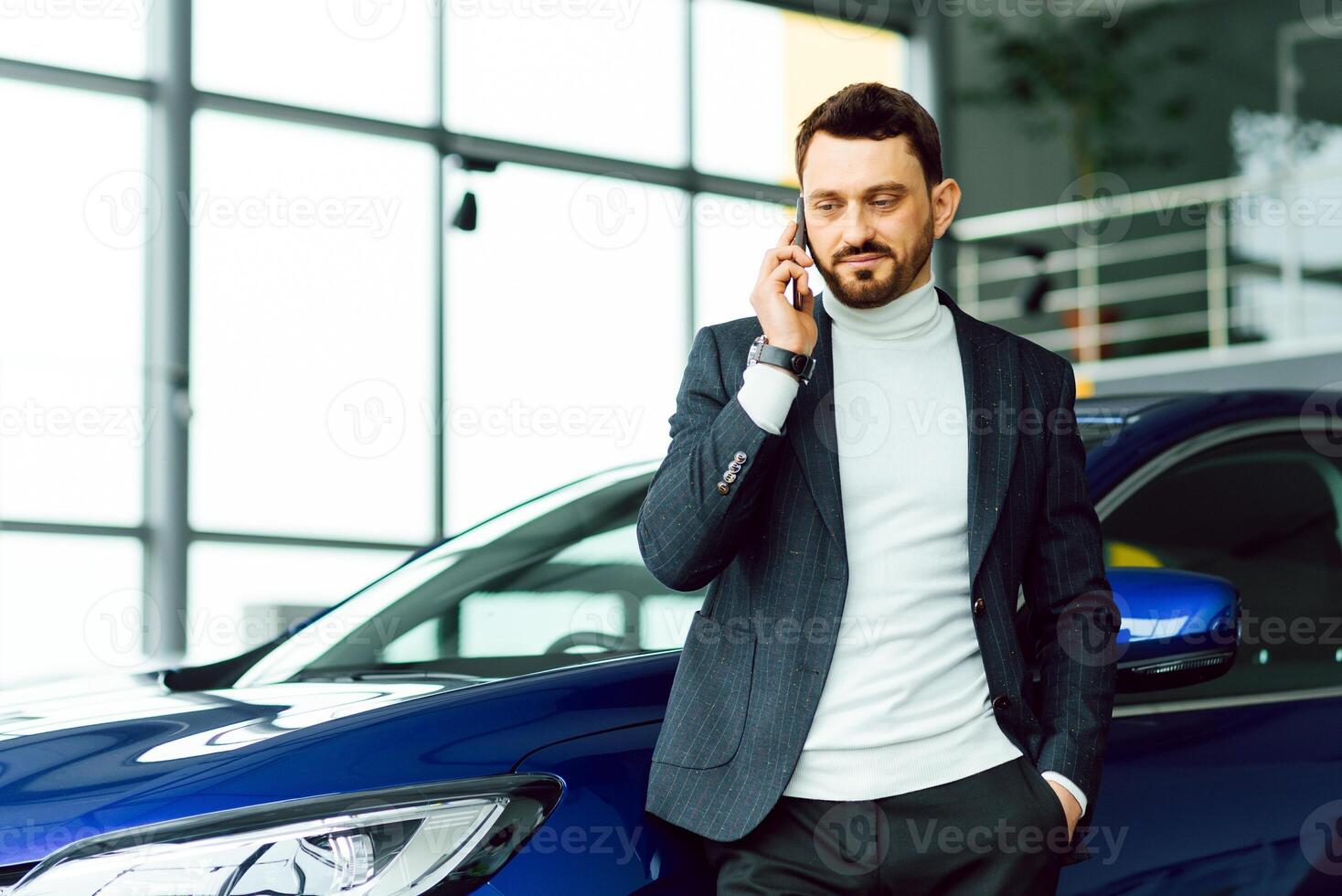 The car shows the character of a man. Confident success elegant business man in full suit and with phone in hand standing near new blue car looks away photo