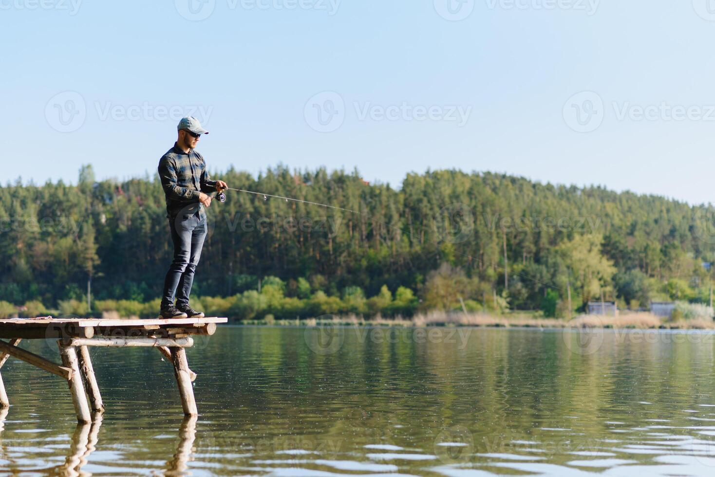 Fishing for pike, perch, carp. Fisherman with rod, spinning reel on river bank. Man catching fish, pulling rod while fishing on lake, pond with text space. Wild nature. The concept of rural getaway. photo