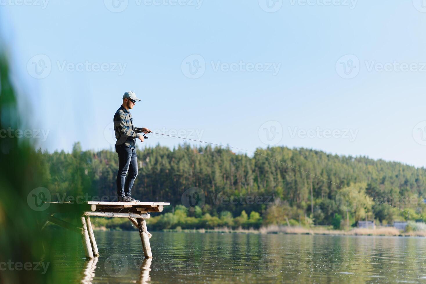 Fishing for pike, perch, carp. Fisherman with rod, spinning reel on river bank. Man catching fish, pulling rod while fishing on lake, pond with text space. Wild nature. The concept of rural getaway. photo