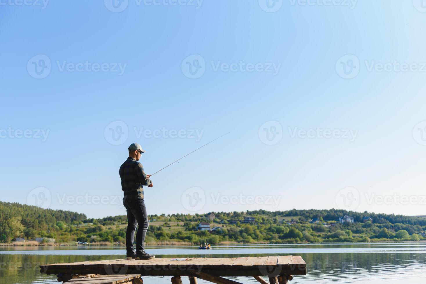 Fishing for pike, perch, carp. Fisherman with rod, spinning reel on river bank. Man catching fish, pulling rod while fishing on lake, pond with text space. Wild nature. The concept of rural getaway. photo