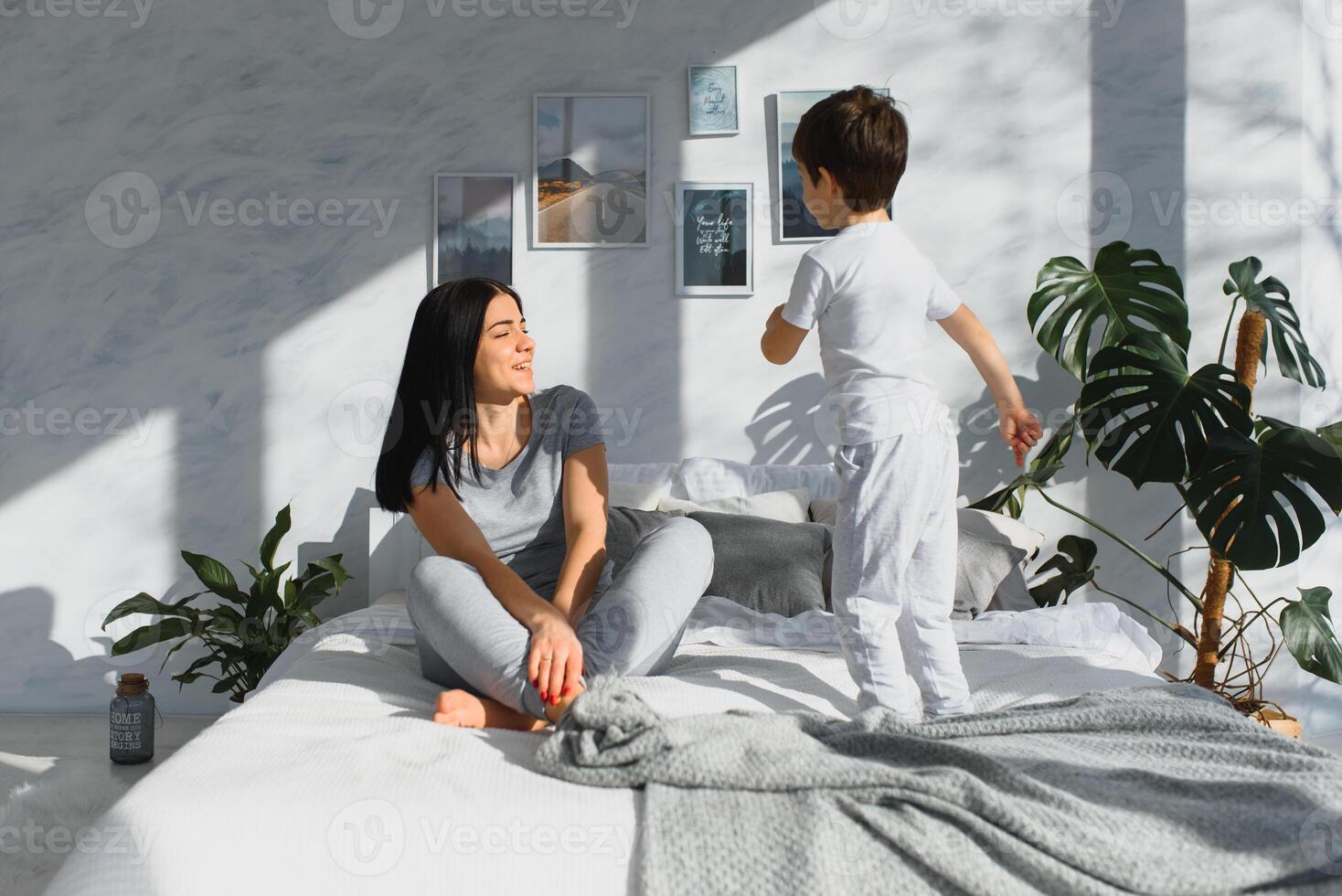 Mom playing with her child in the bedroom. Mother with son sharing good emotions while relaxing at home. Son jumping on the bed. photo