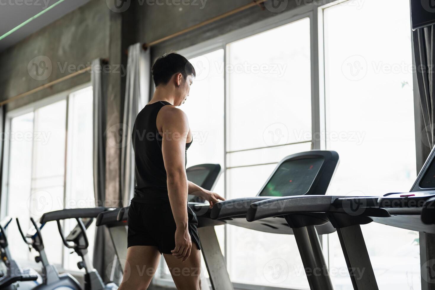 A man in a black tank top sets up a treadmill in a modern gym. His focused expression and muscular build highlight his commitment to fitness and cardio training. photo
