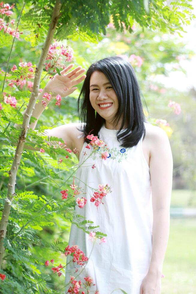 Beautiful asian woman in white dress relax and Sniffing flowers in natural garden. Young Thai girl enjoy on holiday with sunlight in yellow floral park photo