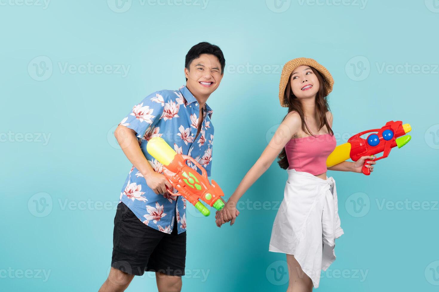 Happy Asian tourist couple in summer clothes using water guns to play Songkran in Thailand. They smile and look at the camera on a blue background with copy space. photo