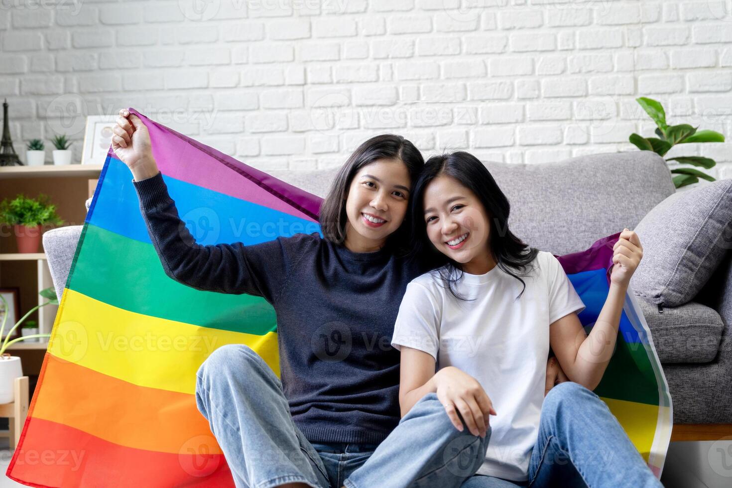Two beautiful young Asian lesbians in casual clothes sitting on the living room floor and smiling together while holding rainbow flags, LGBTQ pride. photo