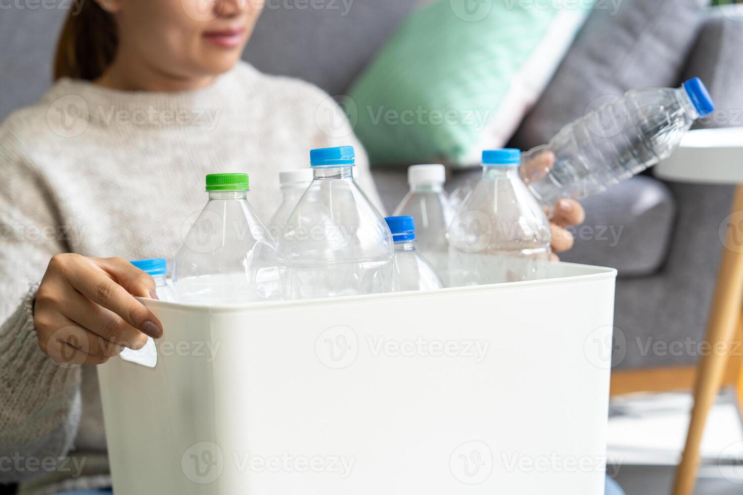 Happy Asian woman wearing a cream-colored sweater collects and sorts recycled plastic bottles into trash bins in the living room. photo