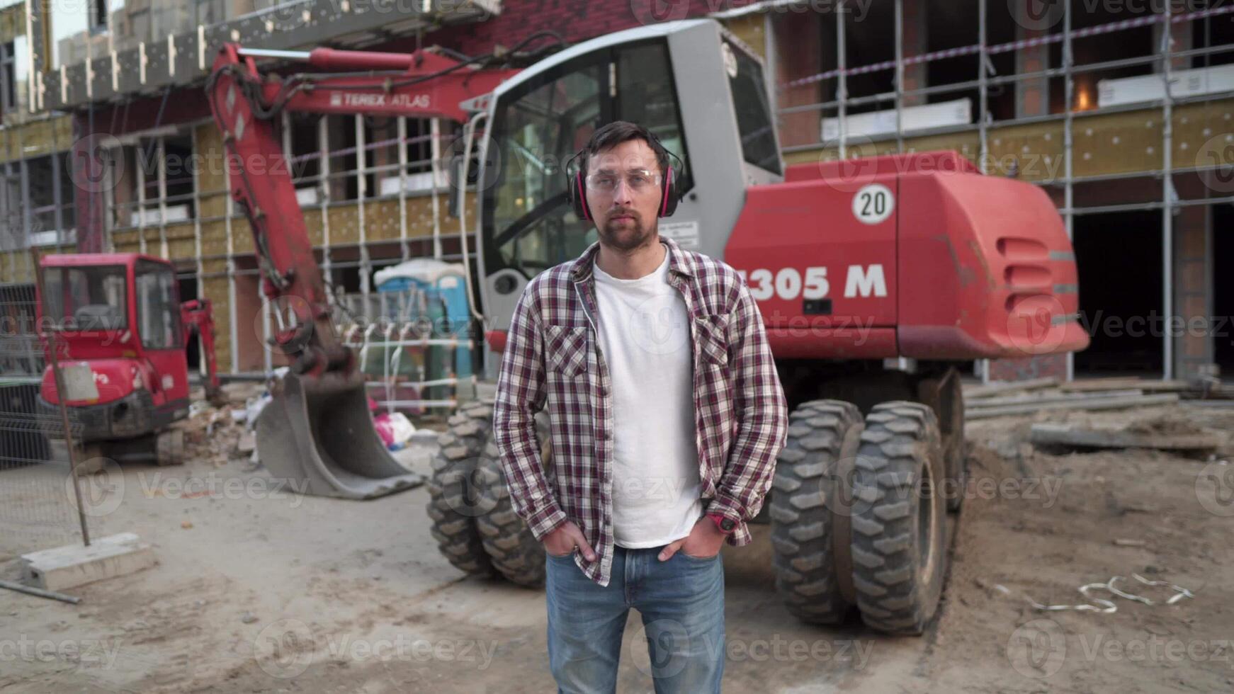 Portrait of successful builder, worker, contractor wearing hard hat and protective earphones, standing on work site with crossed arms confidently. Heavy construction equipment in background. photo