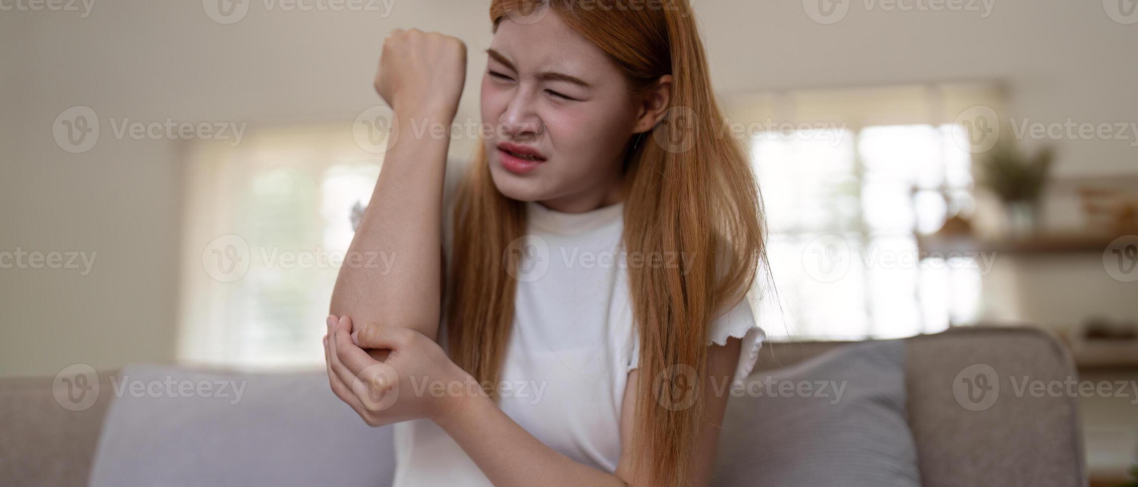 Young Asian woman experiencing severe arm pain while sitting on a sofa at home. Concept of health issues and discomfort photo