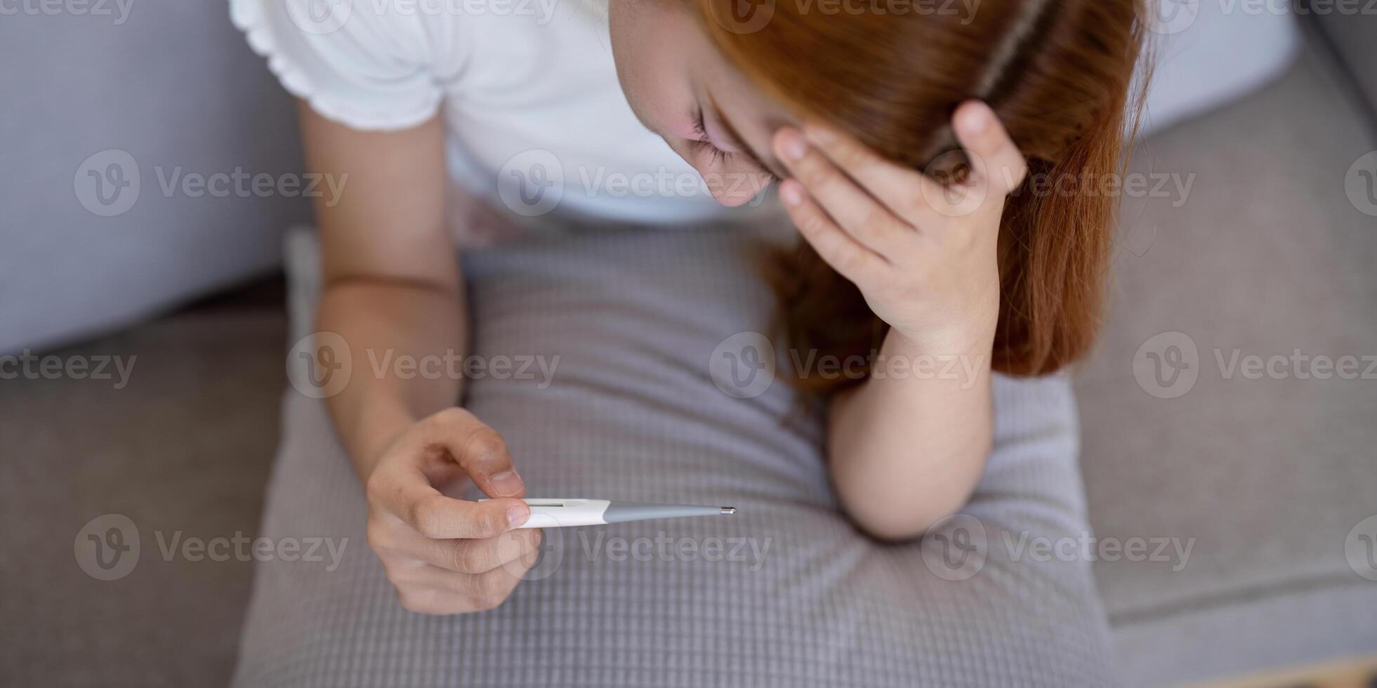 Young Asian woman with fever checking her temperature with a thermometer at home. Concept of illness and healthcare photo