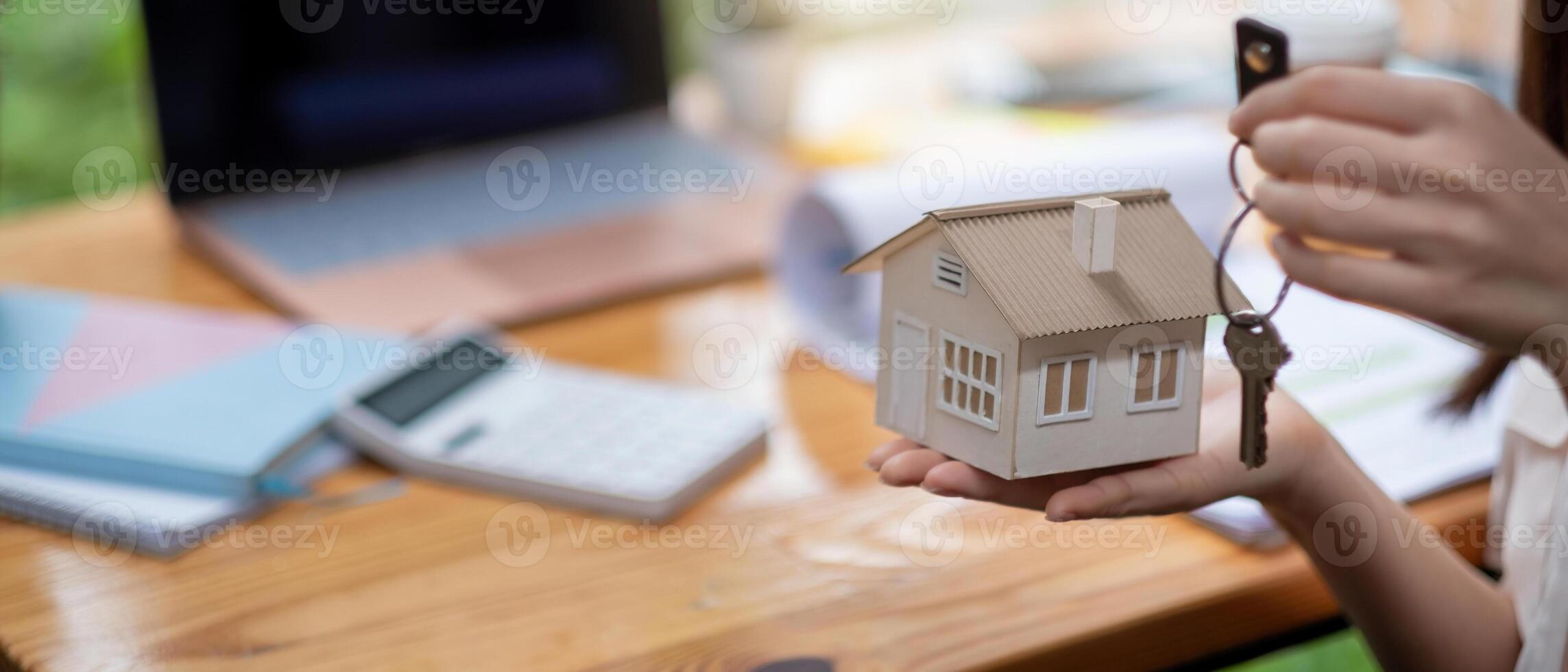 mortgage, real estate and property concept - close up of hands holding house model and home keys with financial working space background. photo
