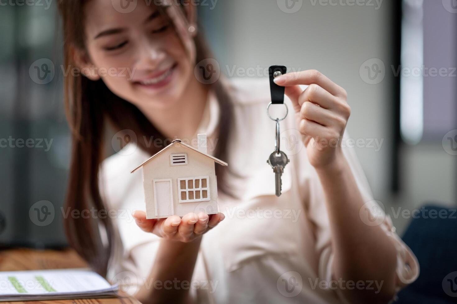 mortgage, real estate and property concept - close up of hands holding house model and home keys at modern office photo