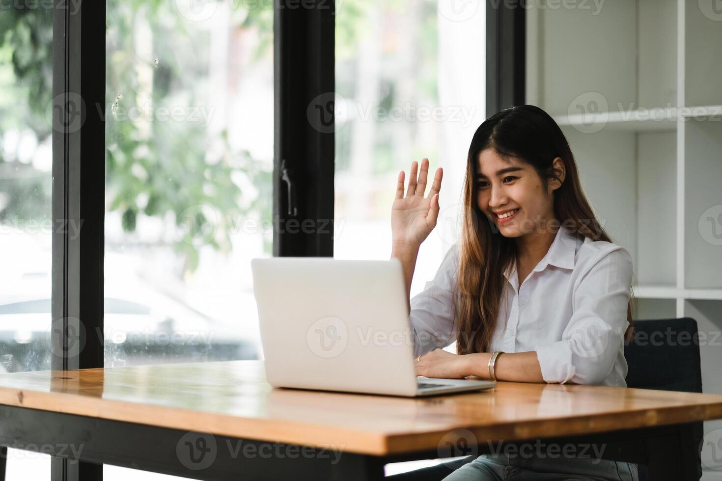 Smiling asian girl talking and waving hand hello during online call via laptop computer, sitting in coffee shop. Cheerful business woman having conference in internet via portable notebook. photo