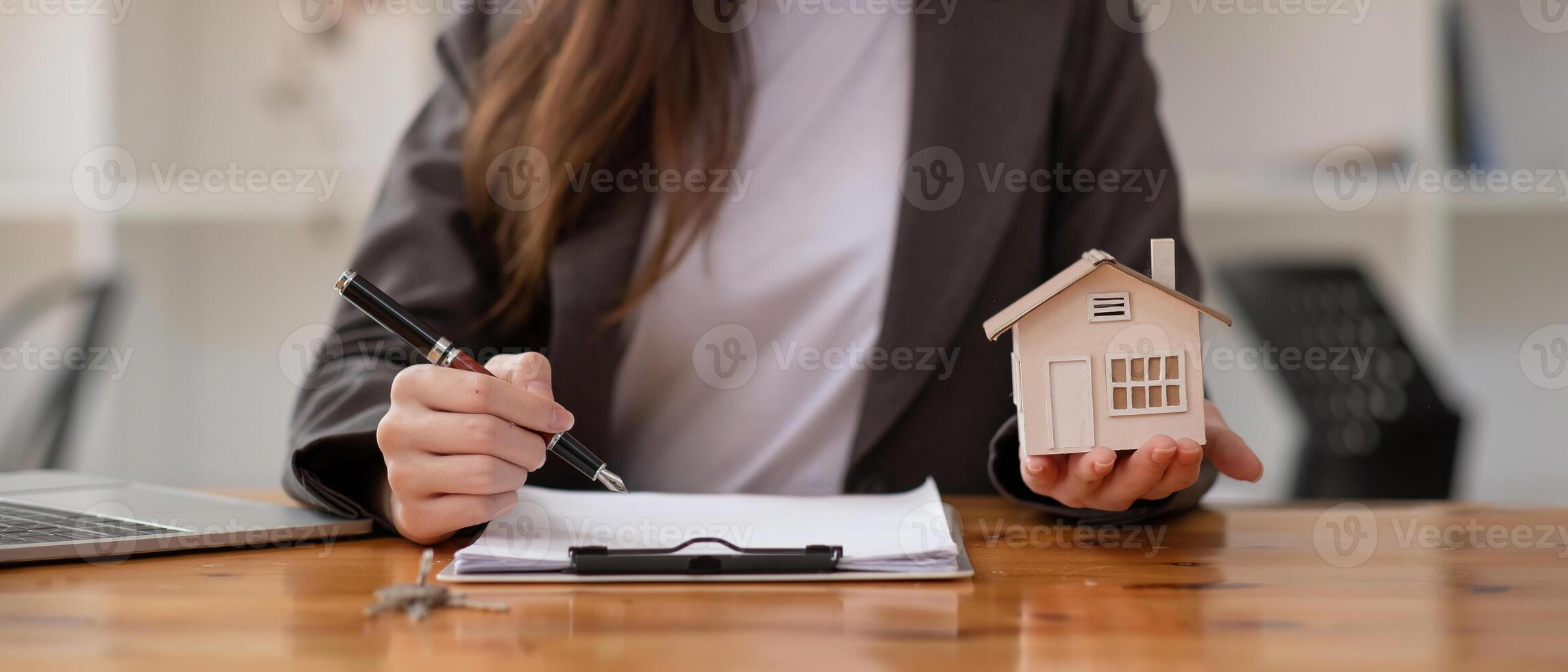 Close up businesswoman realtor taking notes and holding house model, sitting at desk with paper house model and keys, female real estate agent manager, preparing documents, mortgage and property. photo