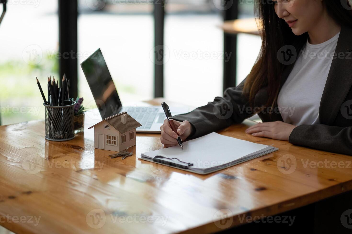 Close up house model miniature on realtor table, businesswoman real estate agent broker writing with documents, taking notes, preparing contract for deal with client, mortgage or rent concept photo
