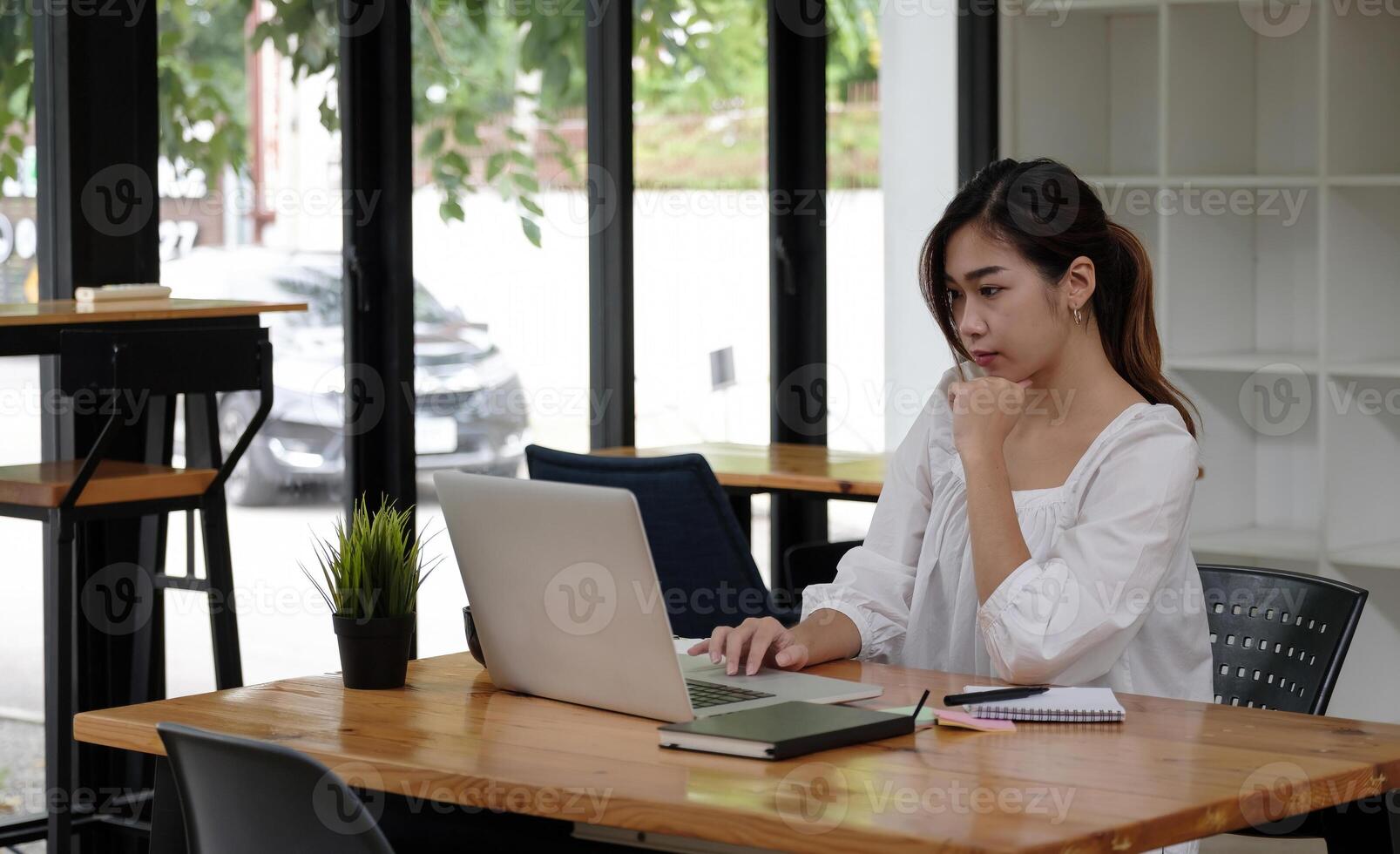 Focused asian business woman working studying online in office looking at laptop, serious employee or student watching webinar writing information in notebook sitting at desk photo