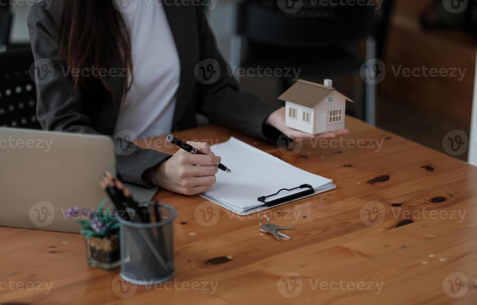 Close up house model miniature on realtor table, businesswoman real estate agent broker writing with documents, taking notes, preparing contract for deal with client, mortgage or rent concept photo