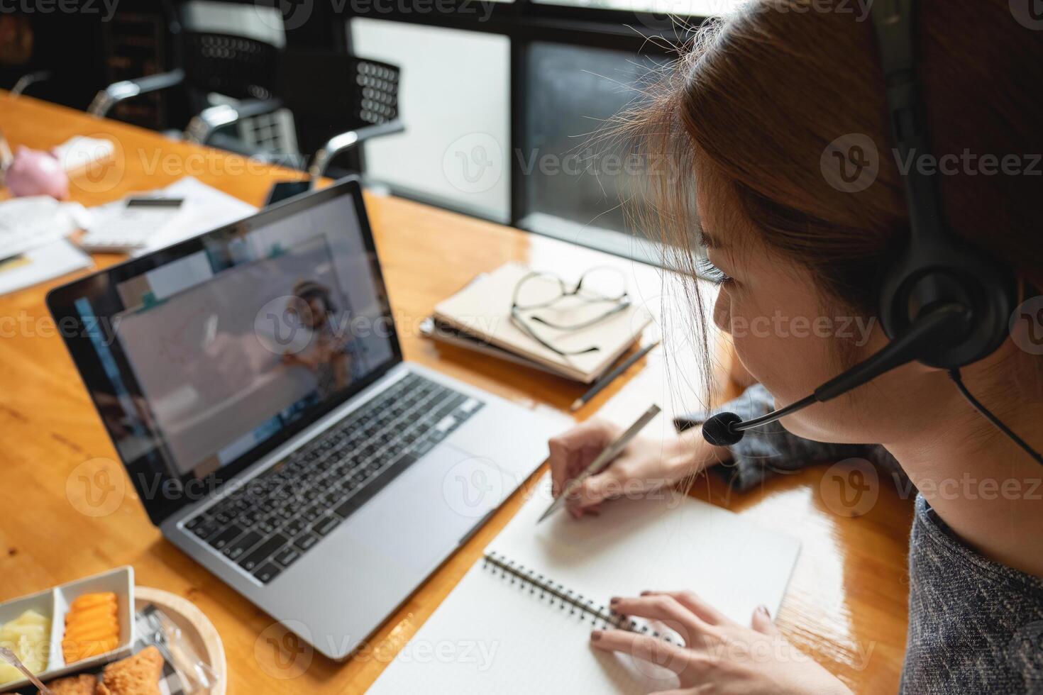 Young student watching lesson online and studying from home. Young woman taking notes while looking at computer screen following professor doing math on call. Girl studying from home on laptop photo