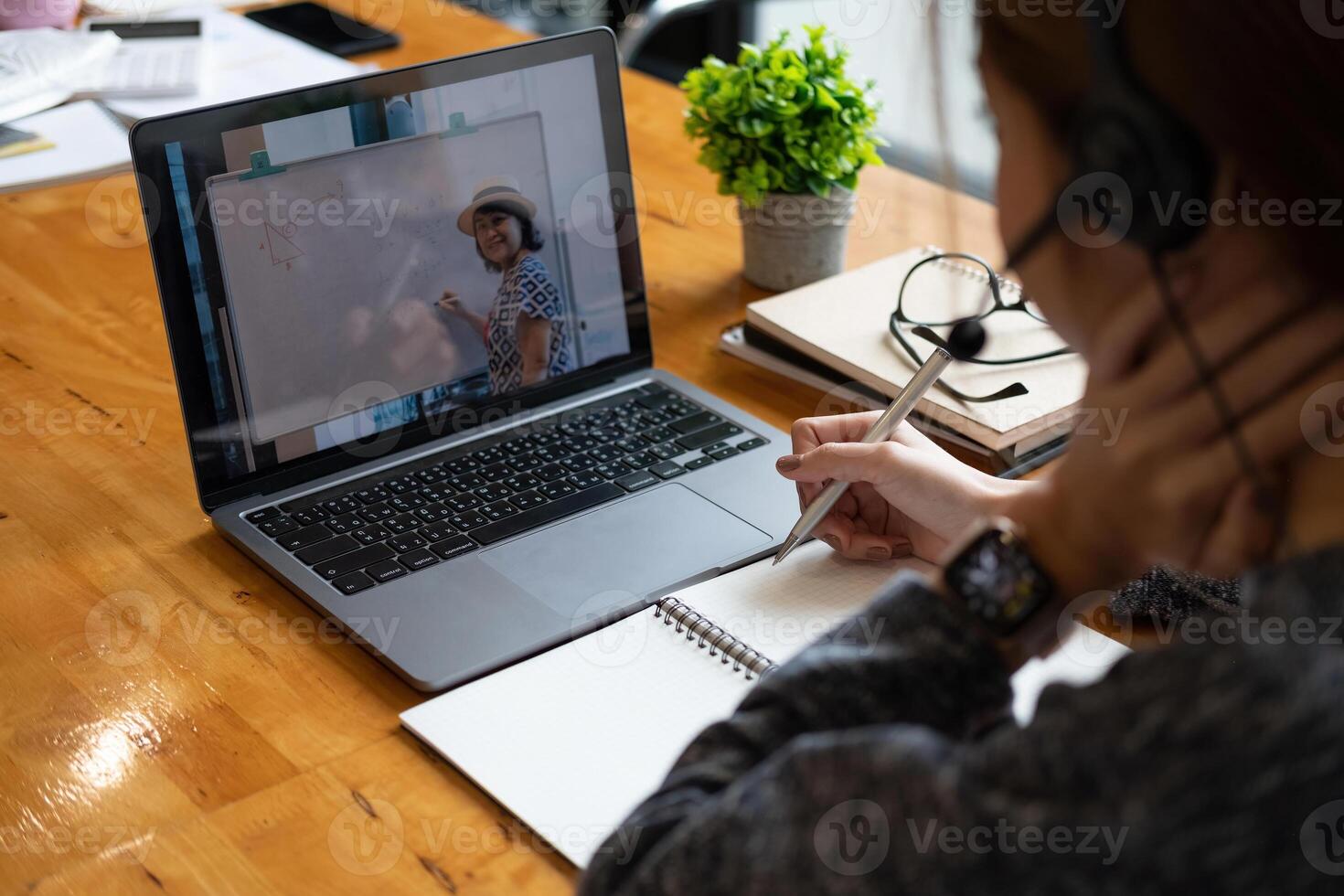 Young student watching lesson online and studying from home. Young woman taking notes while looking at computer screen following professor doing math on call. Girl studying from home on laptop photo