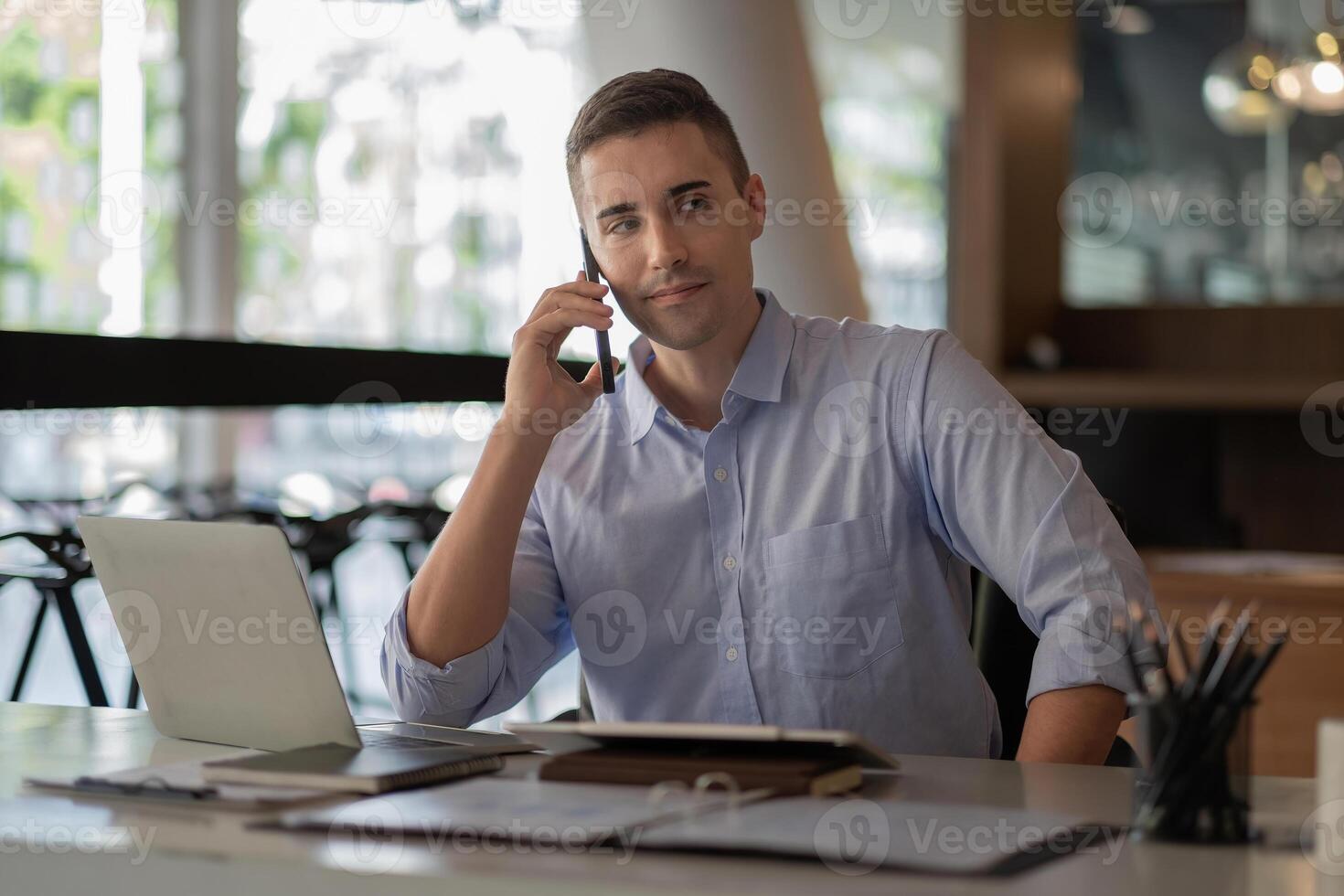 Portrait of young smiling cheerful entrepreneur in modern office making phone call while working with laptop. photo