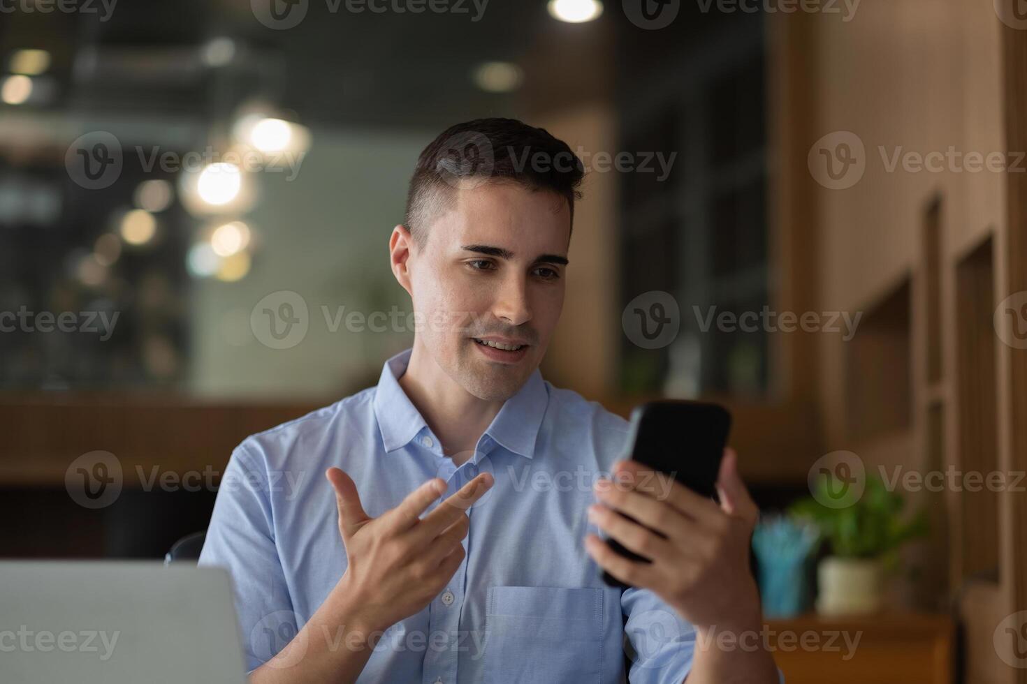 Cheerful happy businessman using smartphone app for virtual communication, holding phone in hand and waving at webcam, holding call sitting at the desk. photo