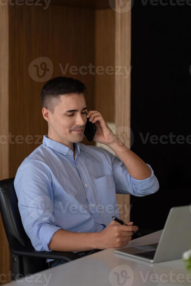 Portrait of young smiling cheerful entrepreneur in modern office making phone call while working with laptop. photo