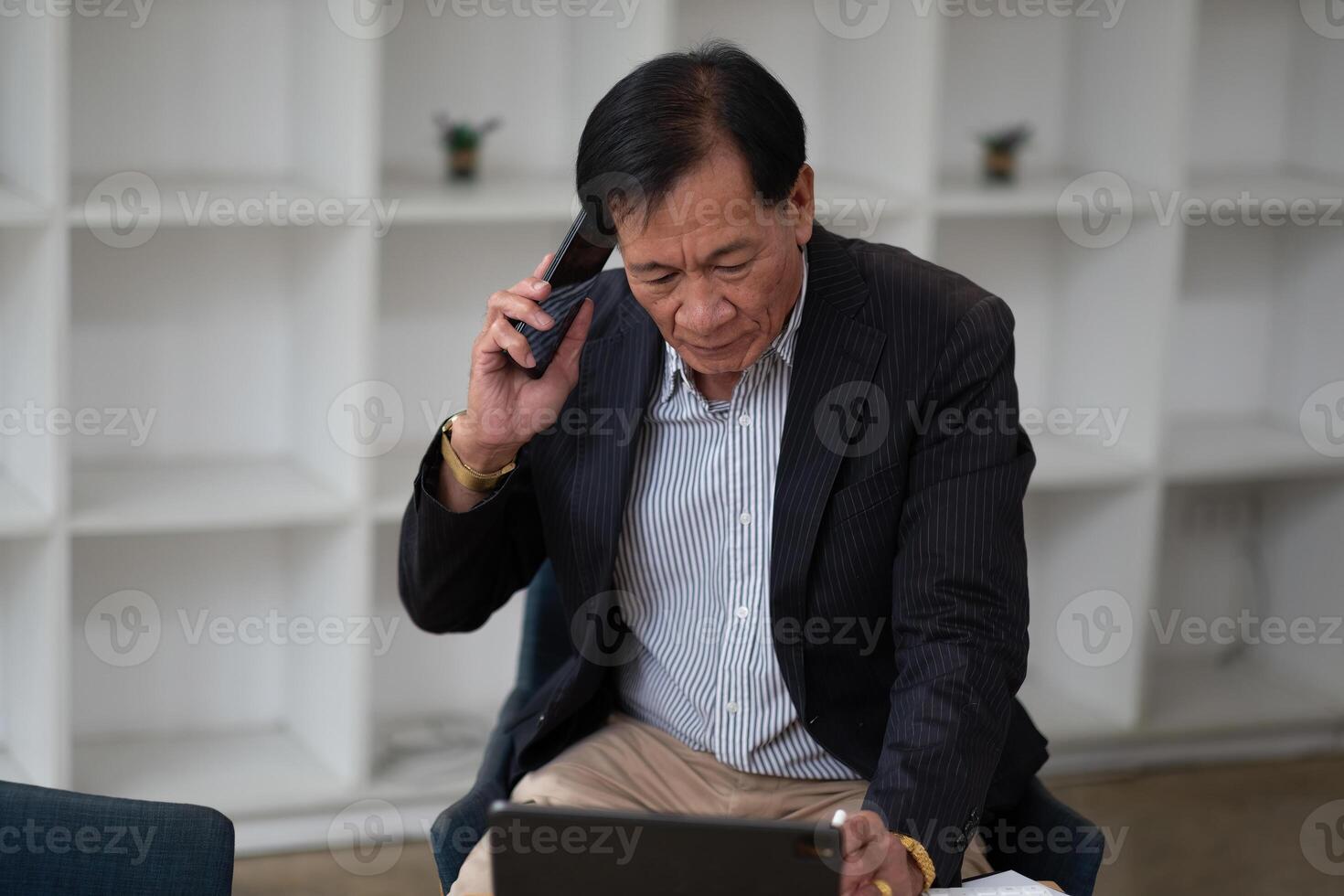 Asian senior manager and businessman in black suit relaxing in social media online and talking, calling with businesspeople on smartphone at workspace in office photo