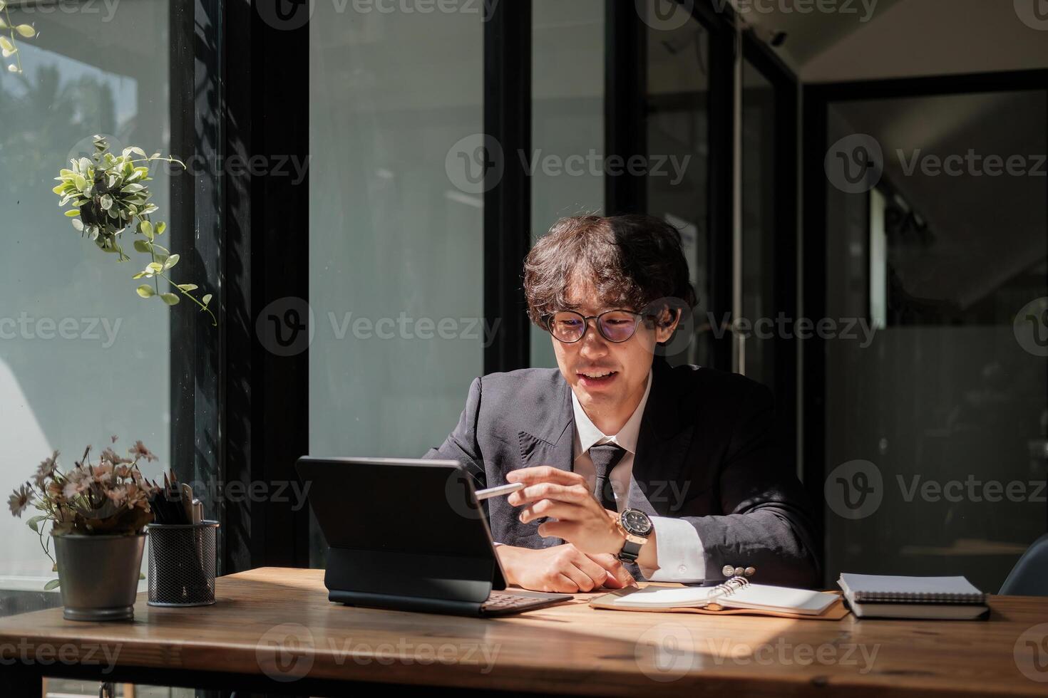 Asian young businessman in eyewear working with digital tablet remotely, sitting at wooden table in cafe. Pleasant happy man communicating in social network, searching information online photo