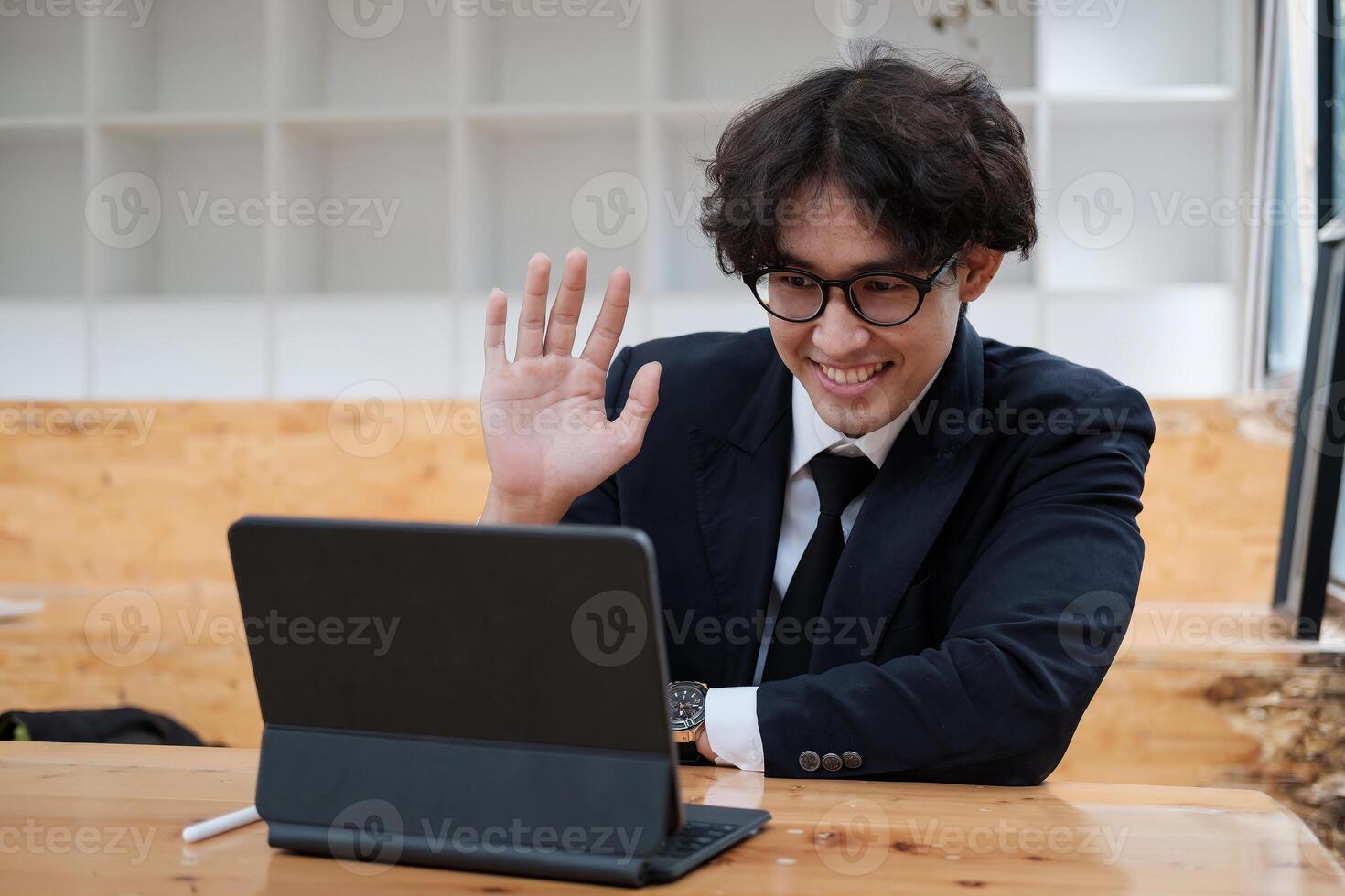 A young asian business man working remotely heaving a meeting on a digital tablet. man waving hand at home during self isolation from coronavirus outbreak crisis photo