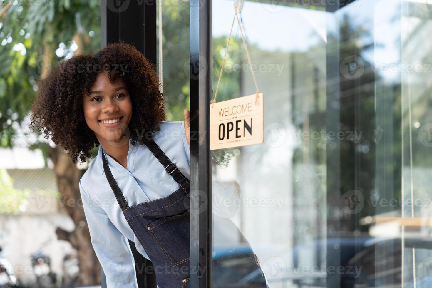 Small business african female owner smiling while turning sign for opening of cafe. Happy afro-american waitress entrepreneur in apron present sign on door photo