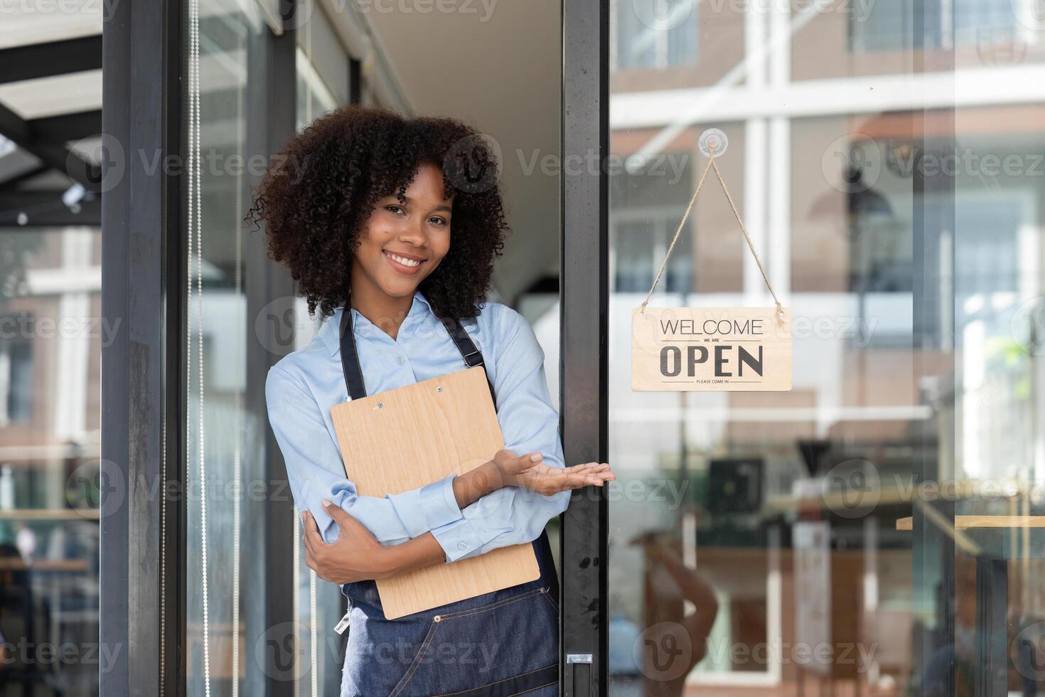 Small business african female owner smiling while turning sign for opening of cafe. Happy afro-american waitress entrepreneur in apron present sign on door photo