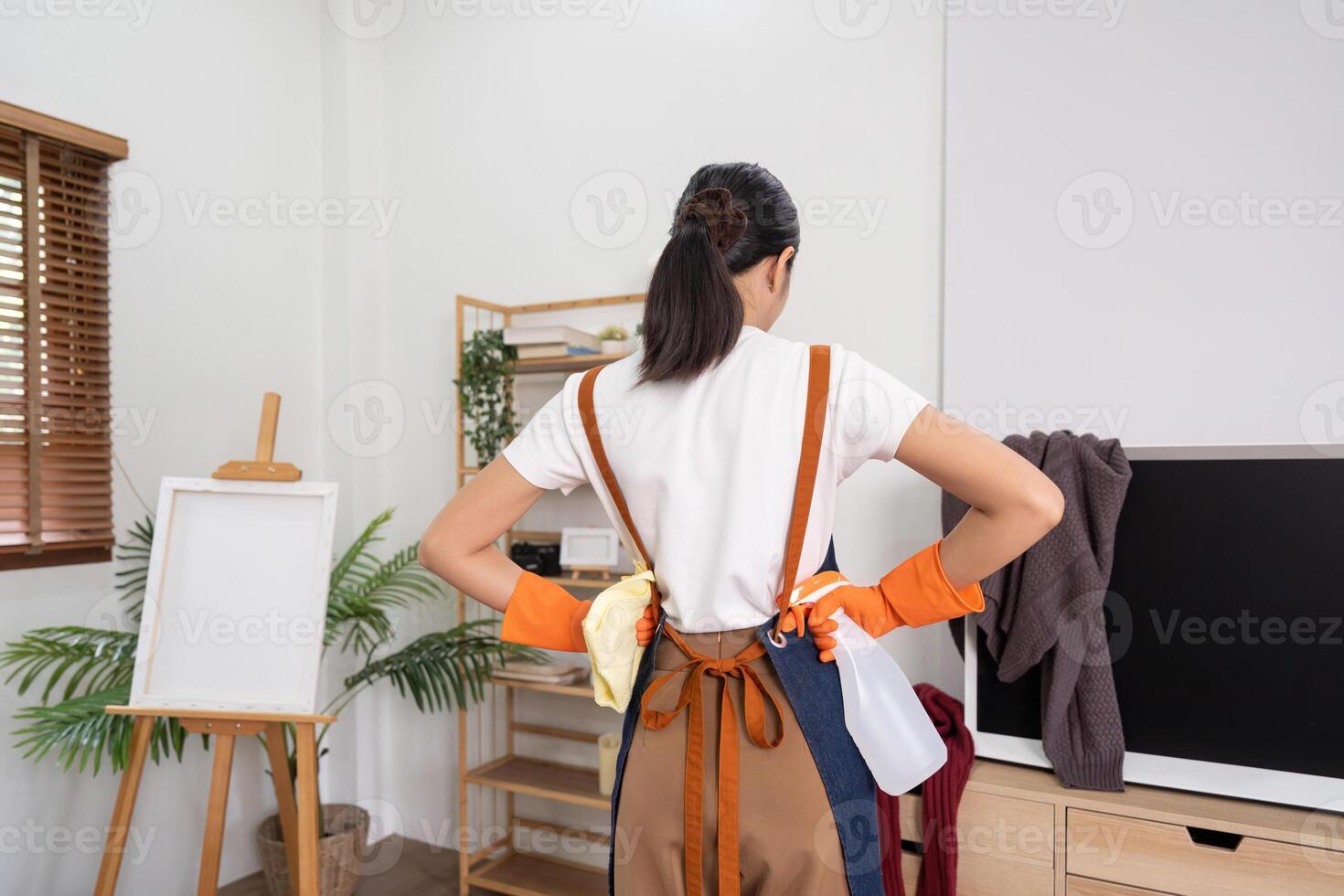 Tired household clean up, housekeeper asian young woman stand and look many clothes on television, cleaning in living room at home. Mess maid or housewife organize dirty and untidy photo