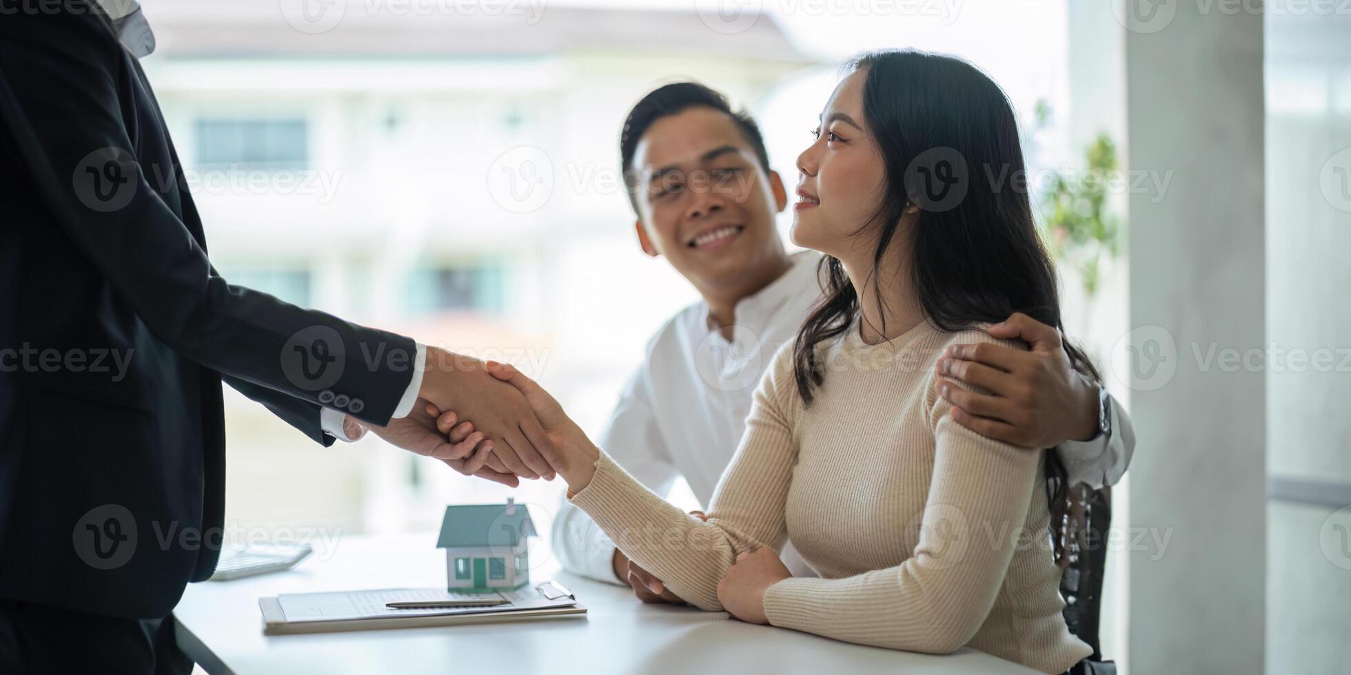Young Asian couple making contract with house sale agency. man and his wife sitting signing the contract next to him looking the contract document with smile. real estate agreement successful concept photo