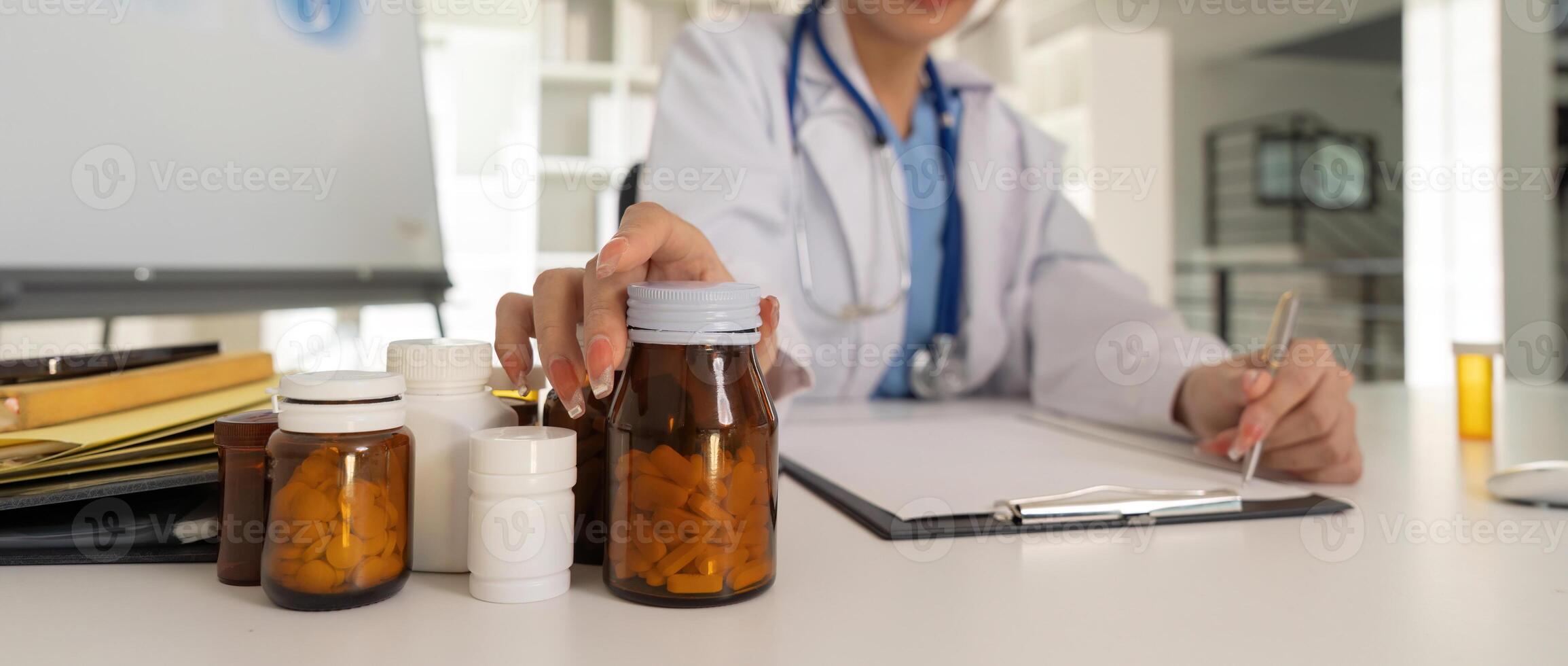 Female doctor writing in a notebook and holding a medicine bottle works at a computer while there were many medicine bottles on the table while sitting at a work desk photo