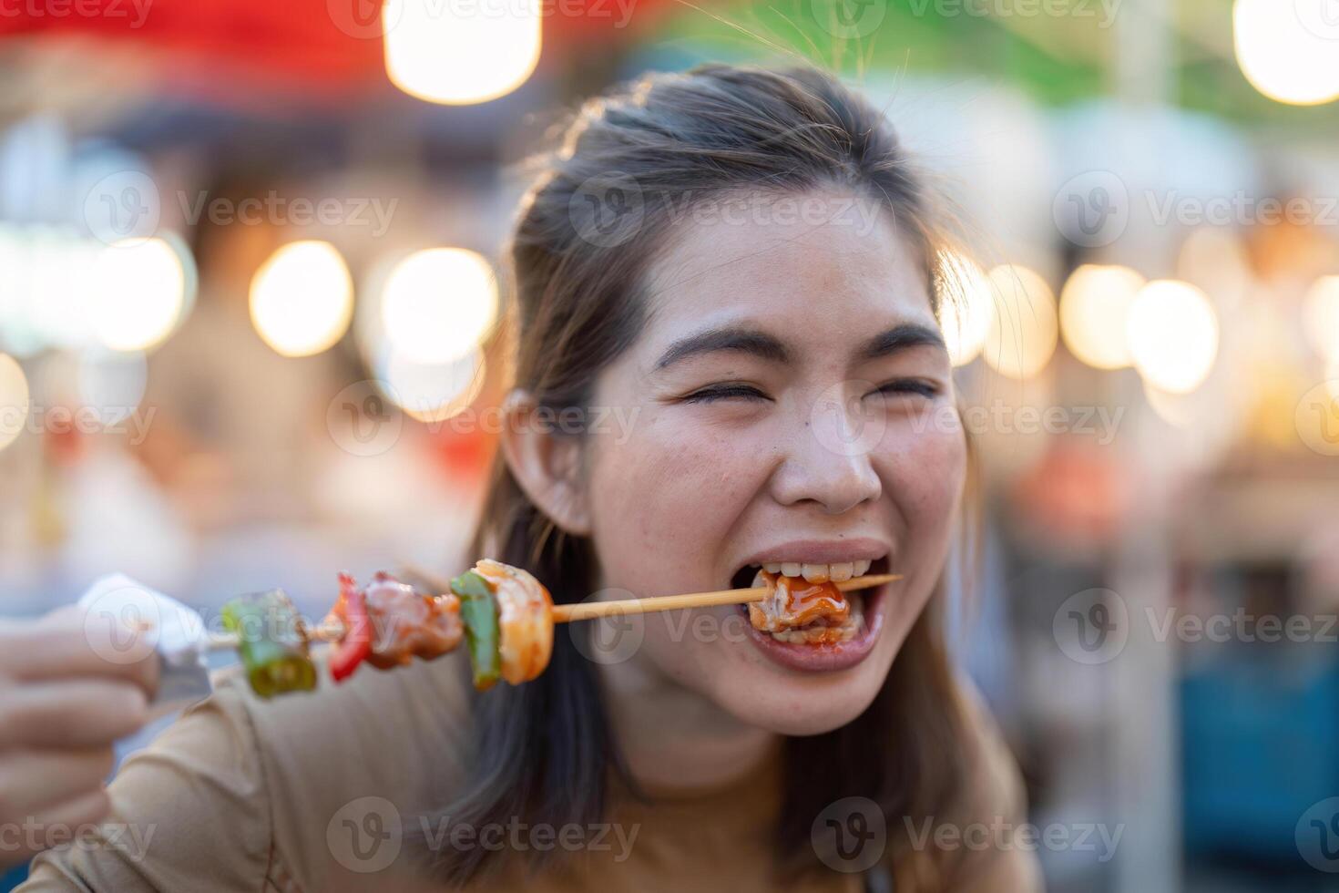 Happy young Asian Traveler foodie woman enjoy eating bbq grilled skewers at outdoor night market street food vendor photo