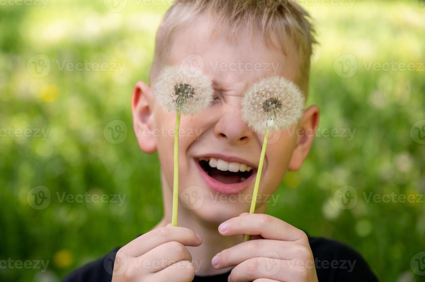 The boy has covered his eyes with dandelions and is smiling widely against a green background. Selective focus. photo