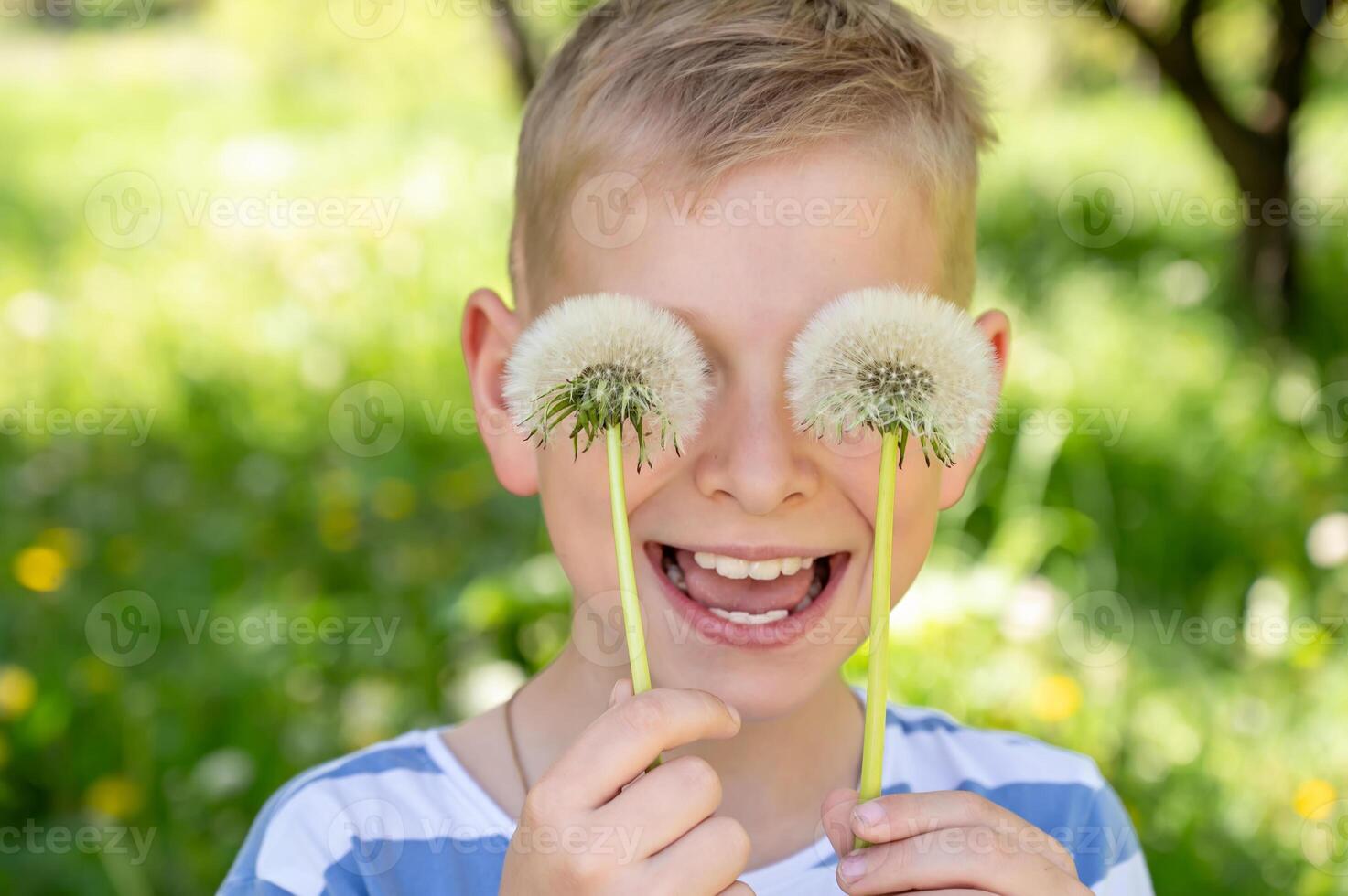 The boy has covered his eyes with dandelions and is smiling widely against a green background. Selective focus. photo