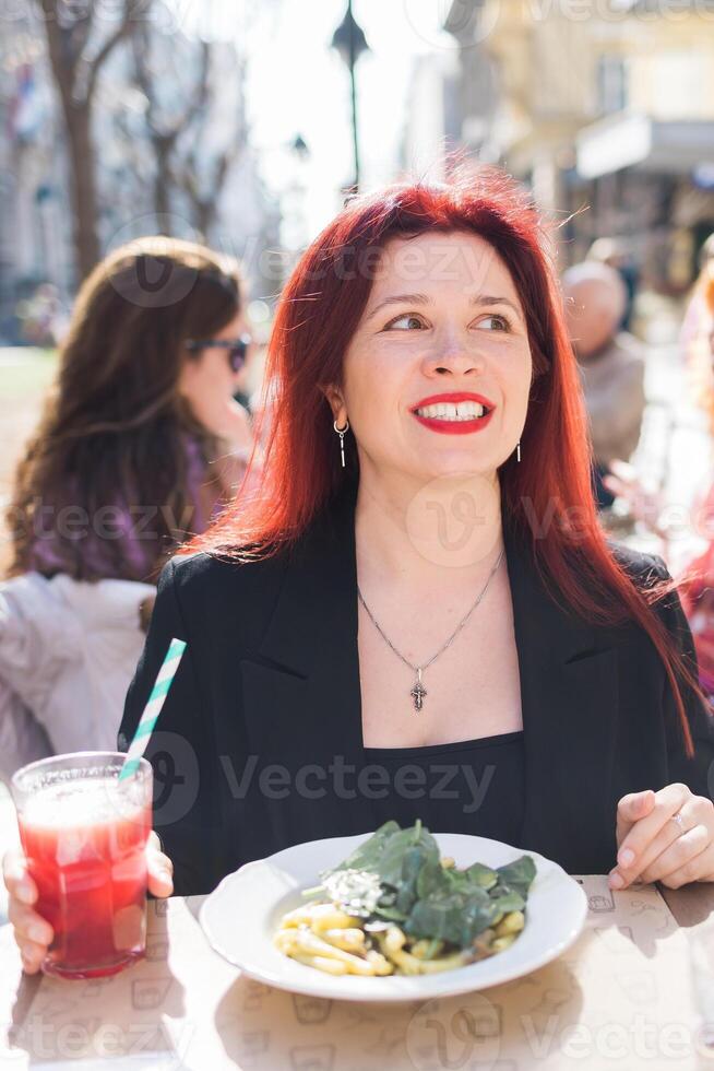 Millennial woman eating italian pasta at restaurant on the street in spring. Concept of Italian gastronomy and travel. Stylish woman with red hair photo