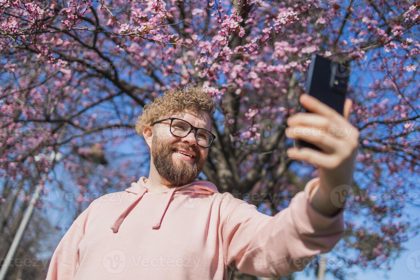 Happy curly man takes selfie against backdrop of flowering tree in spring for his internet communications. Weekend and social networks concept photo