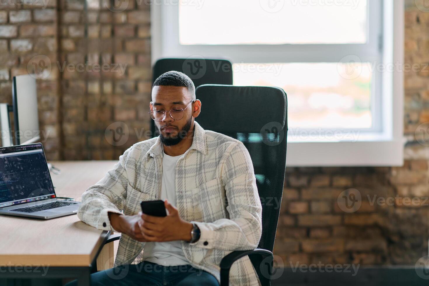 African American entrepreneur takes a break in a modern office, using a smartphone to browse social media, capturing a moment of digital connectivity and relaxation amidst his business endeavors. photo