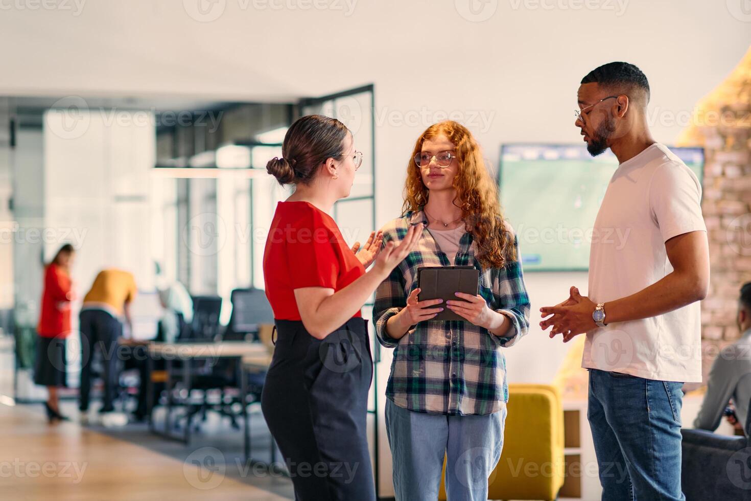 A group of young business individuals, including a girl with orange hair and an African American man, stands in a modern corporate hallway, collectively examining business progress on a smartphone photo