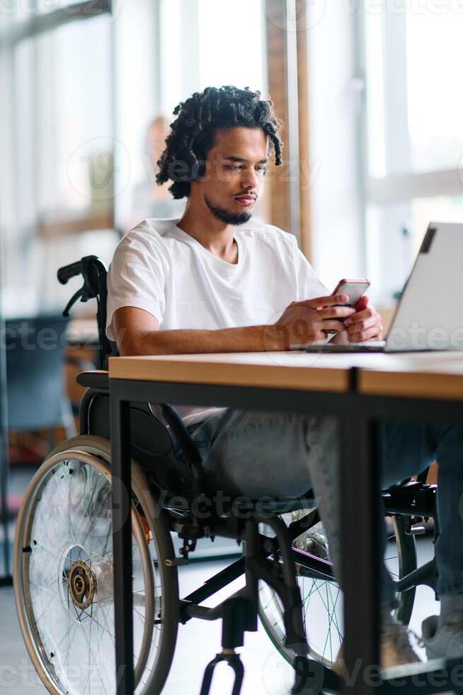 An African American businessman in a wheelchair takes a work break, using his smartphone while seated in a modern business startup coworking center, reflecting both inclusion and technology photo