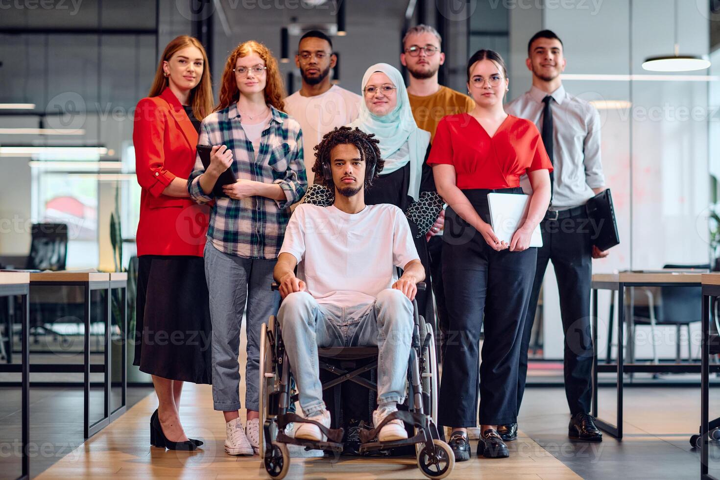 A diverse group of young business people walking a corridor in the glass-enclosed office of a modern startup, including a person in a wheelchair and a woman wearing a hijab photo