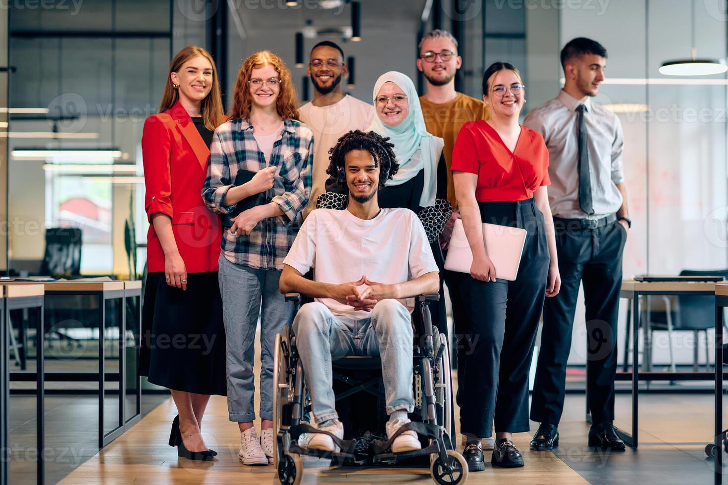 A diverse group of young business people walking a corridor in the glass-enclosed office of a modern startup, including a person in a wheelchair and a woman wearing a hijab photo