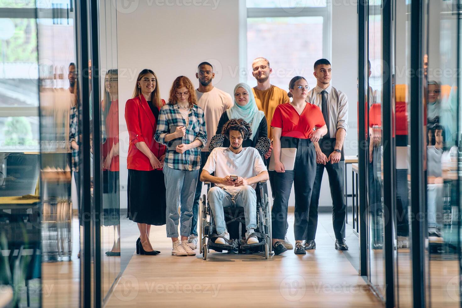 A diverse group of young business people congregates within a modern startup's glass-enclosed office, featuring inclusivity with a person in a wheelchair, an African American young man, and a hijab photo